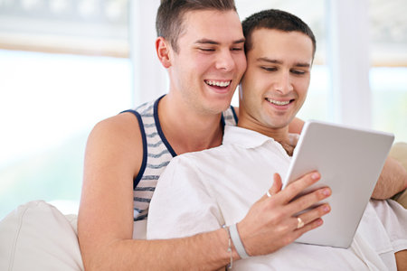 Updating our status as Committed. a young gay couple relaxing on the sofa at home with a digital tablet.の写真素材