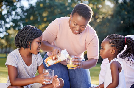 Black woman, park and children family drinks juice for healthy nutrition or wellness nutritionist diet on picnic. Mother with girl kids or black people smile with citrus fruit orange juice in summerの写真素材