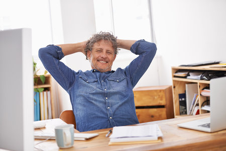 I deserve a breather. Portrait of a mature businessman sitting with his hands behind his head at his desk.の写真素材