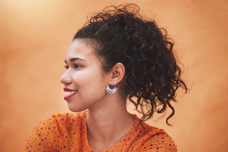 Face, beauty and hair with a woman in studio against an orange background while posing with a smile from the side. Fashion, happy and idea with a young female standing on a bright color wall indoorの写真素材