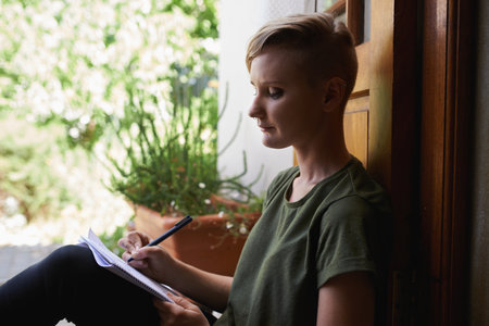 an attractive young woman sitting outside on the porch writing in a notepad.