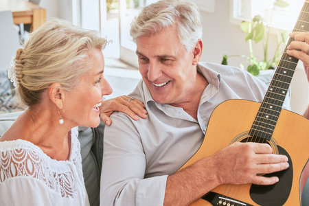 Senior couple with guitar for music and singing together on sofa for retirement lifestyle and summer lens flare. Happy elderly woman or creative musician people in living room home playing love songの写真素材
