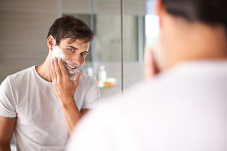 Protecting his face with the correct shaving foam. a young man looking in a mirror to apply shaving foam.の写真素材