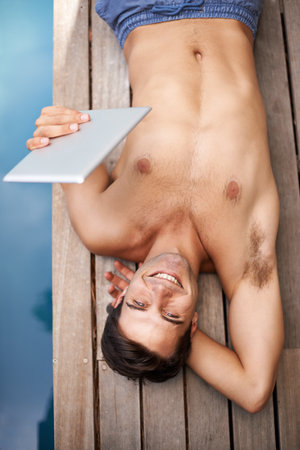 Summer technology. Portrait of a young man lying by the poolside while holding his digital tablet.の写真素材
