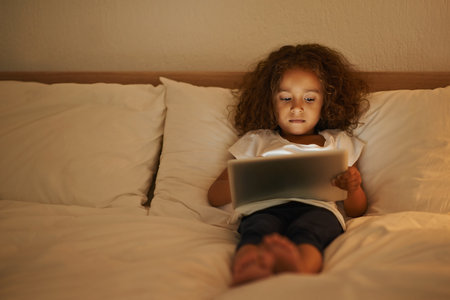 A cute little girl lying in a double bed and playing on a tablet.の写真素材