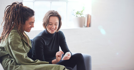 Two women friends using smartphone sitting on sofa at home browsing internet watching online entertainment on mobile phoneの写真素材
