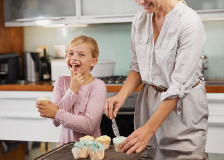 Quick while she isnt looking. Portrait of an adorable little girl helping her mom frost cupcakes in the kitchen.の写真素材