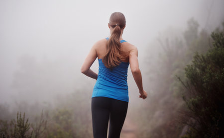 Taking on the trail. Rearview shot of a woman running on a trail on a misty morning.の写真素材