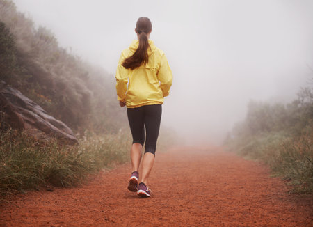 Early morning run. Rearview shot of a woman running on a trail on a misty morning.の写真素材