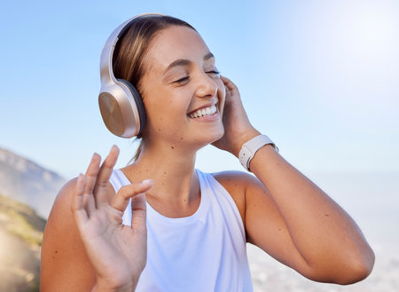 Music streaming, fitness headphones and web radio of a woman athlete listening to a song in nature. Happy smile after a workout on the mountain with blue sky and happiness relax hearing a trackの写真素材