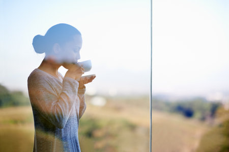 Enjoying a warm cup of coffee while surrounded by nature. Thorugh the window shot of a young woman drinking coffee while standing outdoors.の写真素材