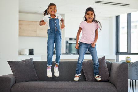 Excited girl kids jumping on living room sofa furniture at home for fun, energy and play games. Portrait crazy, happy and wild young children adhd, action and funny laughing bounce on lounge couchの写真素材