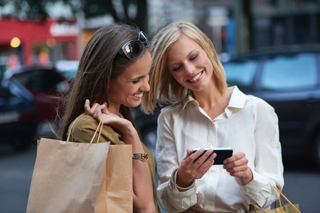 Do you think I should get one of these. A woman showing her friend something on a cellphone while theyre out shopping in the city.の写真素材