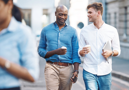 Men, walk and street with takeaway coffee while talk, smile and relax at lunch break. Black man, colleague and diversity in city at tea time, walking to office or workplace in discussion or talkingの写真素材