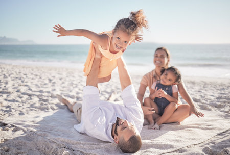 Family, children and playing with a girl on a beach holiday with her parents and sister during summer. Kids, travel and ocean with a female child or daughter on the sand by the sea with her fatherの写真素材