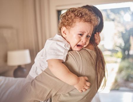 Crying, sad and tears of baby with mom for comfort, safety and attention while hungry, upset or tired in a family home. Toddler boy child cry while in arms of caring woman babysitter, mother or nannyの写真素材