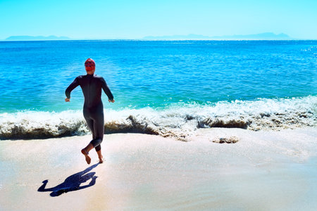 Endurance swimming. Rearview shot of a female swimmer in a wetsuit running towards the surf.の写真素材