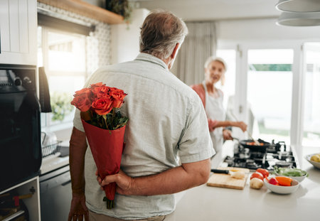 Surprise, roses and a senior couple at home with romantic man spoiling wife with a bouquet of flowers on an anniversary, birthday or valentines day at home. Husband hiding gift for a woman behind hisの写真素材