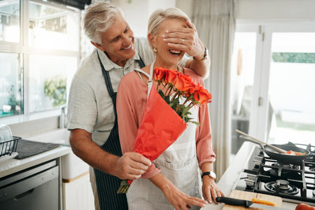 Senior couple, covering eyes and flowers surprise as man give wife bouquet of roses on an anniversary, birthday or valentines day in kitchen. Happy old man and woman being romantic in Australia houseの写真素材