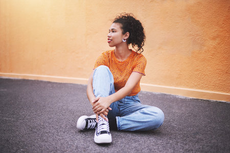 Fashion, sneakers and young woman on street in the city. Portrait of urban, trendy and black female model against an orange wall background sitting on the road. Style, clothes and girl posingの写真素材