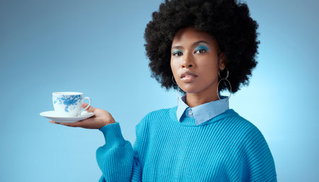 Tea, fashion and blue with a black woman in makeup on a wall background in studio holding a cup and saucer. Face, portrait and afro with a young female posing for drink, beverage and refreshmentの写真素材
