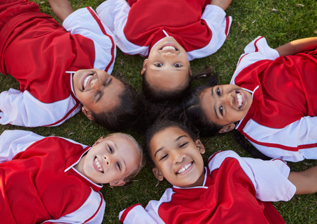 Happy football children relax on field sports portrait for group collaboration, teamwork and motivation. Soccer girl kids face lying on ground together for training support, love and commitment aboveの写真素材