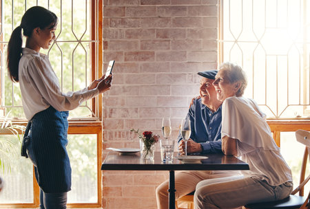 Photograph, restaurant and date with a senior couple in celebration of an anniversary and waitress using a phone. Service, romance and travel with a man and woman tourist posing for a pictureの写真素材