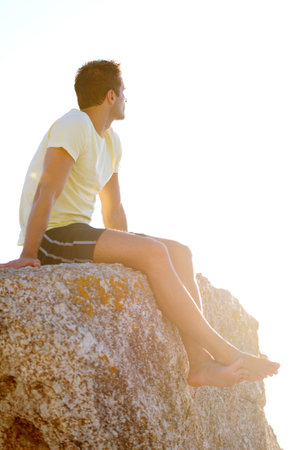 Enjoying the view. Young man sitting on a rock with the sunset in the background.の写真素材