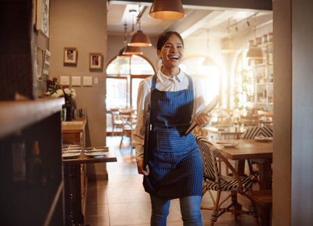 Woman, happy and waitress in restaurant working in apron with food menu in hand for table. Girl, smile and service work at luxury diner, cafe or coffee shop show happiness on face for job in Londonの写真素材