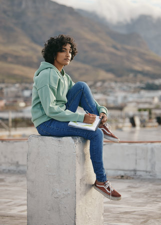 Art, city and a woman drawing in notebook sitting on rooftop looking for inspiration and creativity from cityscape. Creative girl from India on roof, thinking and relax with sketchbook and pencil.の写真素材