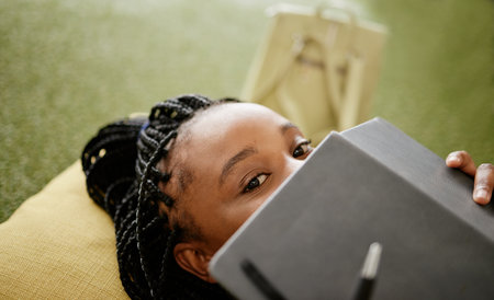Black woman and student hiding face with notebook on casual college study lounge bean bag. Shy African university learner on studying rest break looking with lecture book to hide identity.の写真素材