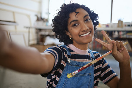 Art selfie, peace sign and woman in studio workshop happy, smile or ready for creative oil painting for artwork design. Artist portrait of India painter after working on drawing with watercolor brushの写真素材