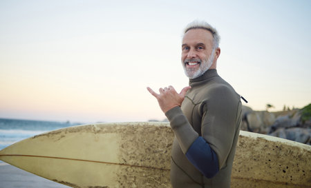 Summer, surfing and portrait of old man at the beach in the morning. Retired senior man with surfboard, hand sign and smile on face ready to surf at sunrise. Ocean, hobby and water sport in Australiaの写真素材