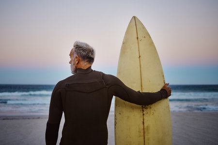 Beach, man and surfboard with back view for morning cardio fitness and tranquil swim in nature. Senior surfer waiting for low tide at ocean for calm surf waves with peaceful sky at dawn.の写真素材