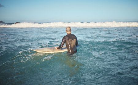 Surfer and man in water watching wave for high tide while holding surfboard at sunny USA beach. Retirement person enjoying surfing sport leisure in California waiting for ocean level to rise.の写真素材