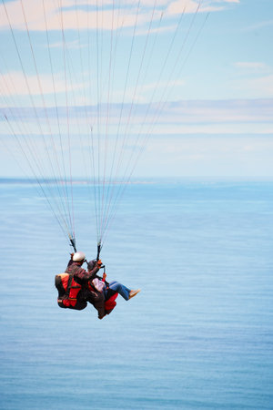 Over the ocean. Side view of two young people doing tandem paragliding with the ocean in the...