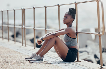 Beach, relax and woman runner thinking after an outdoor cardio workout in nature by ocean. Tired African athlete, running and fitness girl sitting on promenade to breathe, calm and rest after workoutの写真素材