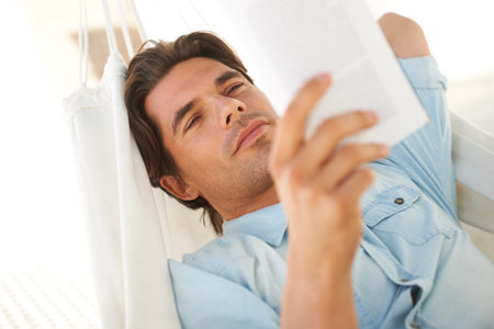 Taking some time for himself. A young man reclining on a hammock while reading a novel.の写真素材