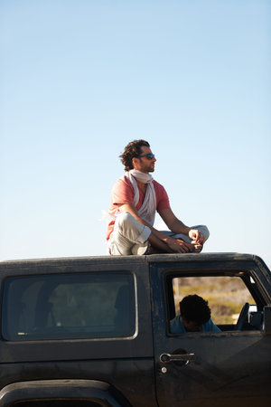 Cooling off outside. A young man sits on the car roof while his friend looks at the map inside.