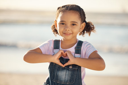 Girl at beach make heart sign with hands, happy and smile against blurred nature background. Young female child with expression of happiness, makes love icon, gesture with fingers by the ocean or seaの写真素材