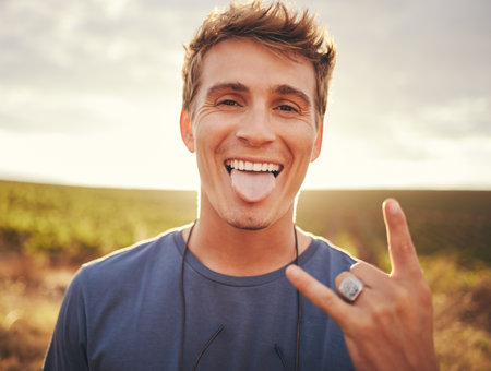 Man, hands and portrait of rock sign for travel tour or vacation in the countryside and nature outdoors. Happy, excited young male traveler, rocker or musician with hand gesture and tongue outの写真素材