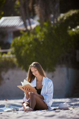 One beautiful young caucasian woman relaxing on the beach. Enjoying a summer vacation or holiday outdoors during summer. Taking time off and getting away from it all. Reading alone on the sand outsidの写真素材