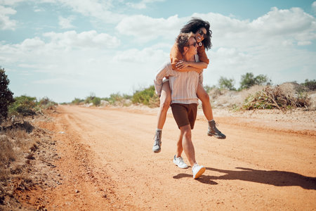 Couple, love and travel with a man and woman walking on a sand road in the dessert or nature together. Summer, romance and dating with a happy, young male giving a piggyback to a female in the wildの写真素材