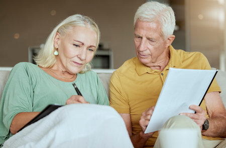 Unhappy elderly couple sitting on a sofa together and looking stressed. Senior caucasian man and woman looking worried about their future while looking at paperwork and their debtの写真素材