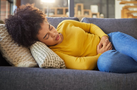 Young unhappy mixed race woman holding her stomach and suffering from stomach pain on the couch at...