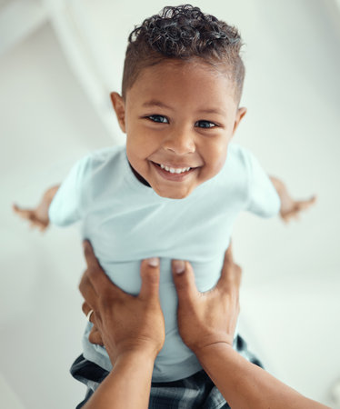 Happy little mixed race boy lying in fathers arms while looking at the camera and smiling. Loving family with father lifting cute little child son playing plane and having fun while bonding at homeの写真素材