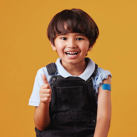 Portrait of a smiling happy mixed race child giving the thumbs up to the vaccine while showing his bandaid and posing against an orange copyspace backgroundの写真素材