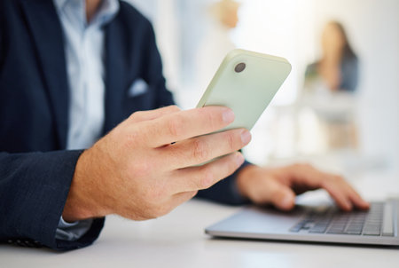 Closeup of one caucasian businessman browsing on a laptop and sending texts on a cellphone in an office. Hands of entrepreneur planning and doing banking on his digital wireless devicesの写真素材