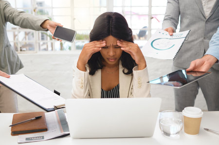 Young african american businesswoman suffering from a headache while using a laptop and working in a demanding environment. Stressed businessperson looking tired while working with a group of colleagの写真素材