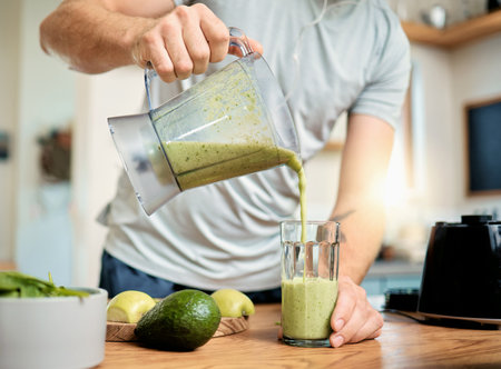Closeup of one caucasian man pouring healthy green detox smoothie from blender into glass in kitchen at home. Guy having fresh fruit juice to cleanse and provide energy for training. Wholesome drinkの写真素材